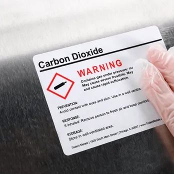 Hand holding carbon dioxide warning label with red diamond hazmat symbol and safety instructions