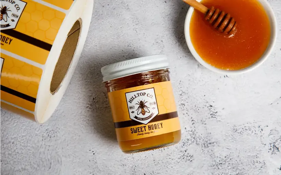 a labeled jar of honey between a roll of honey labels and a bowl of honey with a honey wand