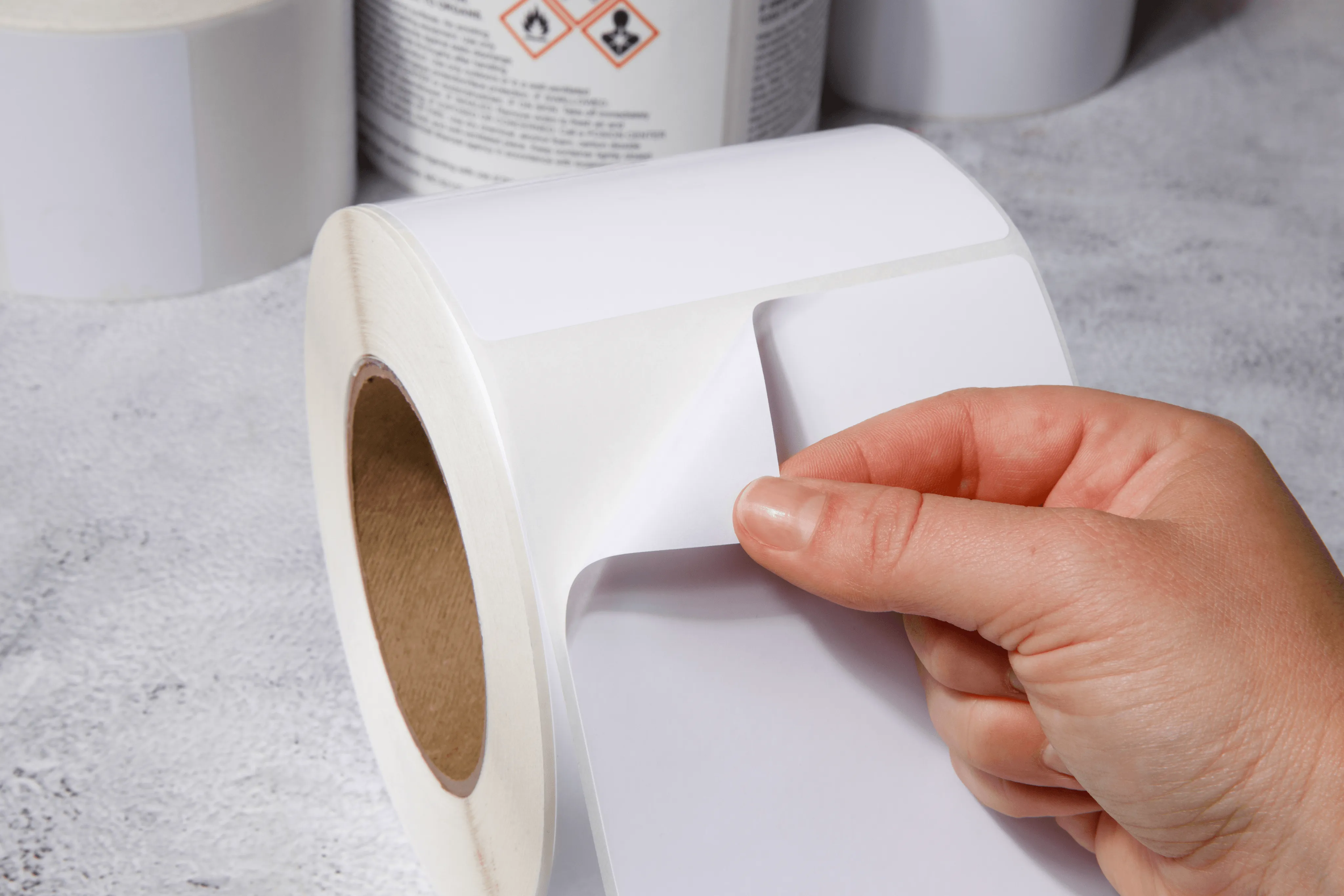 hand holding white thermal label roll with industrial tracking labels and hazmat symbols visible