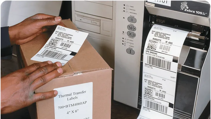 a hand applying a thermal shipping label to a cardboard box with a label printer in the background